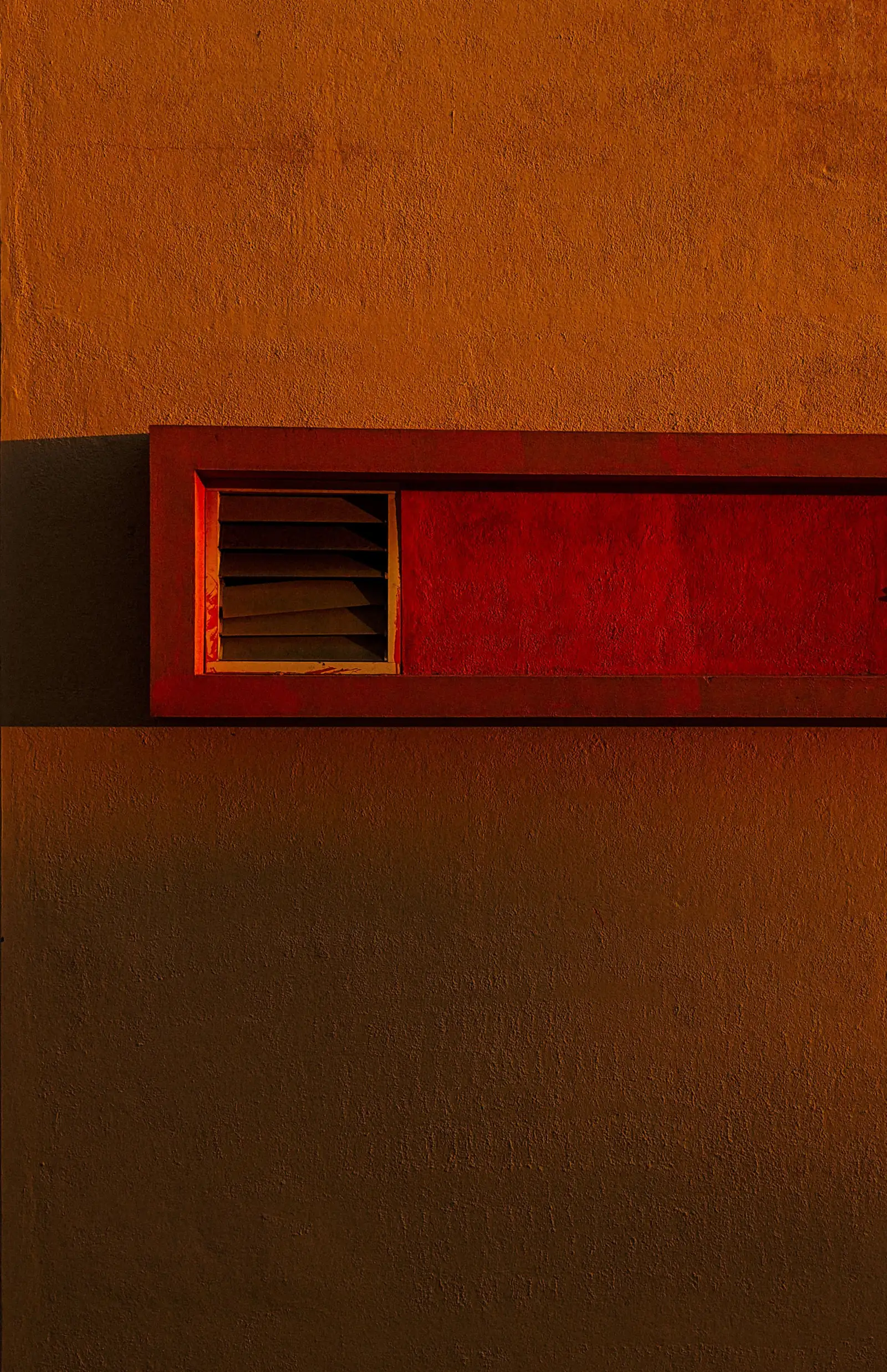 A red-framed louvred vent set into a sunlit ochre wall